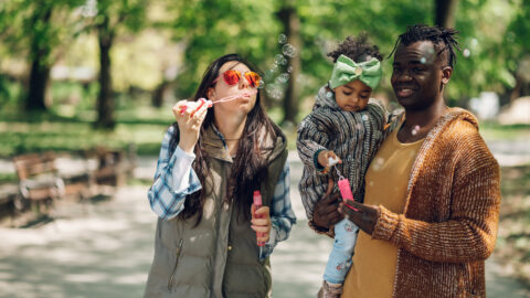 Beautiful baby girl enjoying making soap bubbles in the park while spending a beautiful sunny day with her diverse parents outside. Happy multiracial family concept.